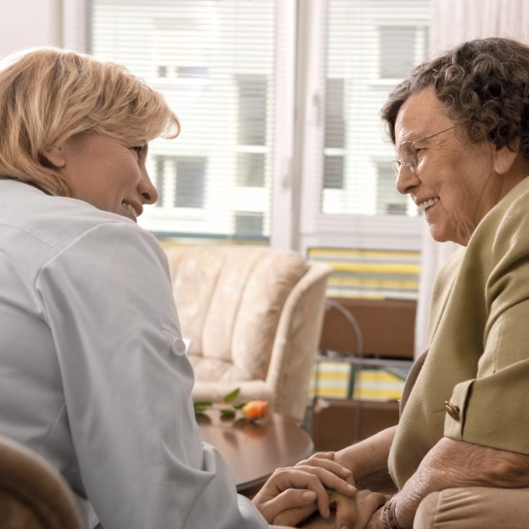 Eine mittelalte Frau sitzt einer Seniorin gegenüber und hält ihre Hände. Beide lächeln.