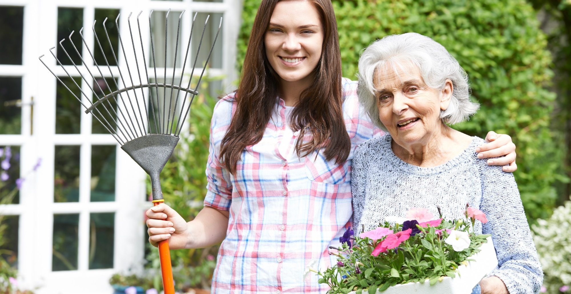 Im Garten ist eine Frau, mit Rechen in der Hand und dem Arm um eine Seniorin gelegt, die eine Kiste mit zu pflanzenden Blumen im Arm hat.